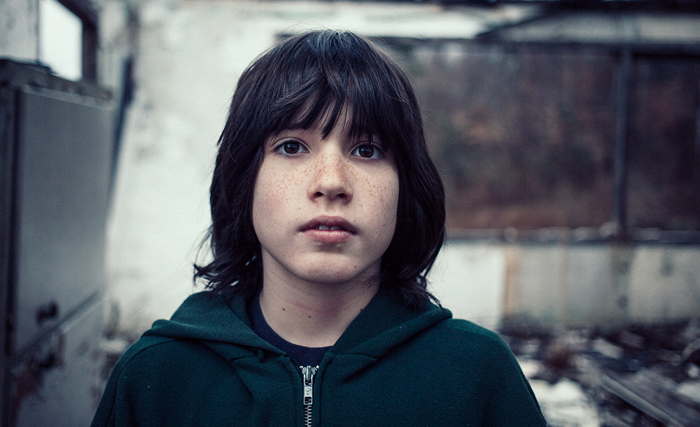 A young person with freckles in a green hoodie inside a derelict building